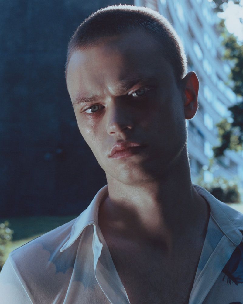 Close-up portrait of a young adult male with short hair wearing an open white collared shirt, captured outdoors with natural lighting.