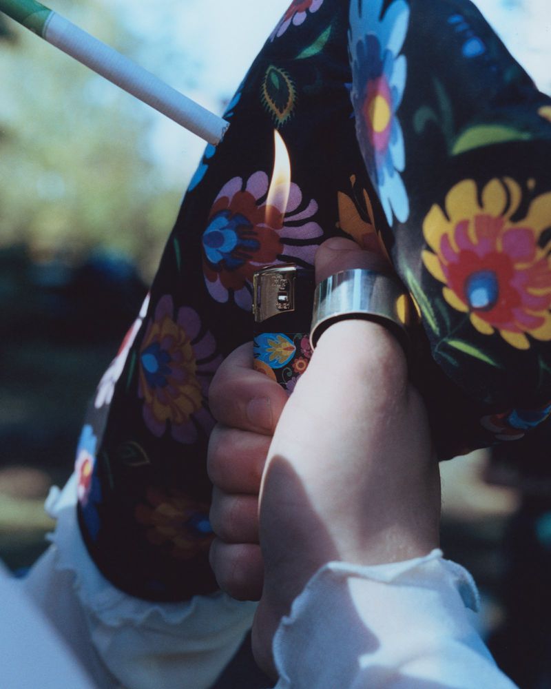 Close-up of hand holding a lighter to a cigarette, featuring a floral print sleeve, silver ring, and neutral background. Smoking concept.
