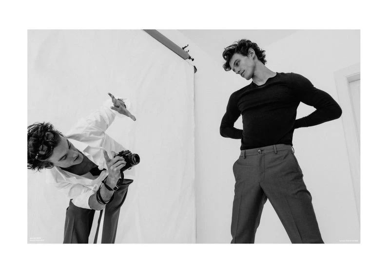 Two men in a photo studio: one adult male with a vintage camera photographing another adult male in a dark shirt and grey pants.