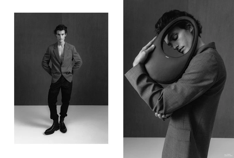 Black and white fashion shot of a male model posing in a blazer and trousers, holding a black handbag in a studio setting.