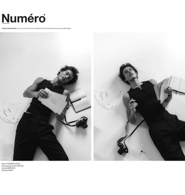 Monochrome fashion editorial shot of a young adult man lying on a white surface, surrounded by papers, with a vintage camera nearby.