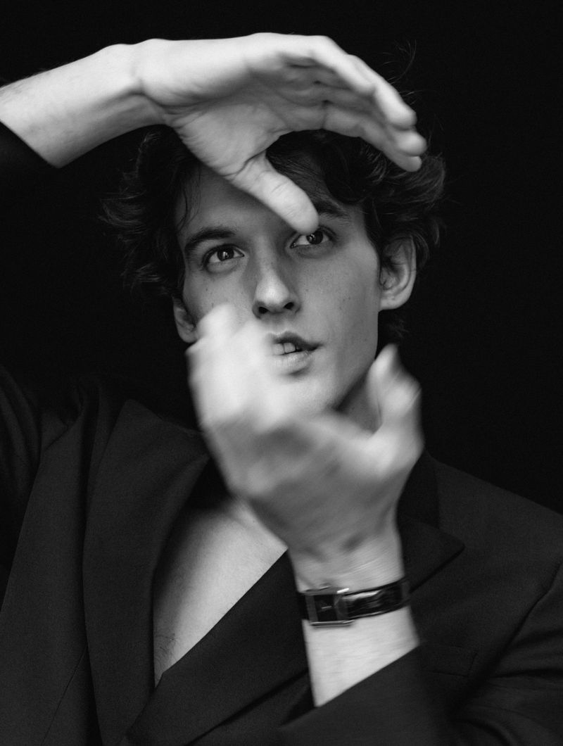 Monochrome portrait of a young adult man with dark hair wearing a blazer, framing his face with his hands against a black background.