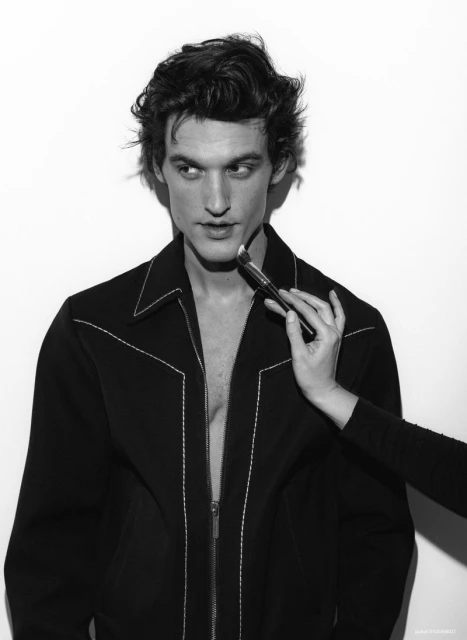 Black and white portrait of a male model having makeup applied with a brush on a white backdrop in a photo studio setting.