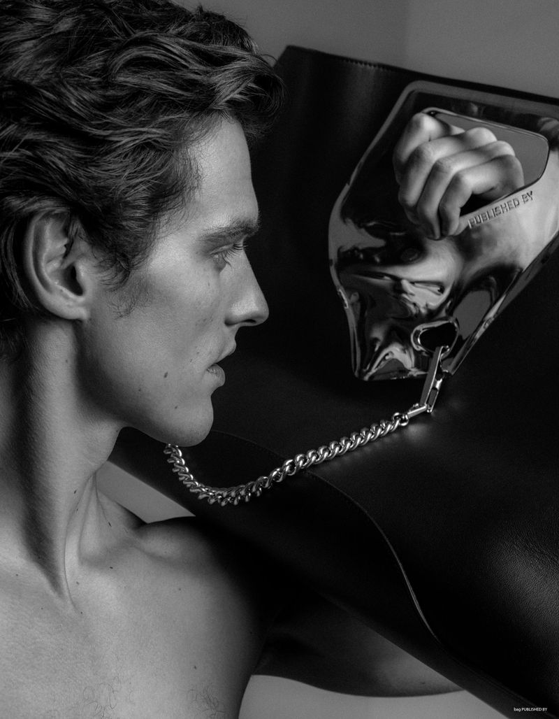 Monochrome image of a shirtless adult male in profile, with dark wavy hair, posing with a large black leather tote bag with a silver handle.