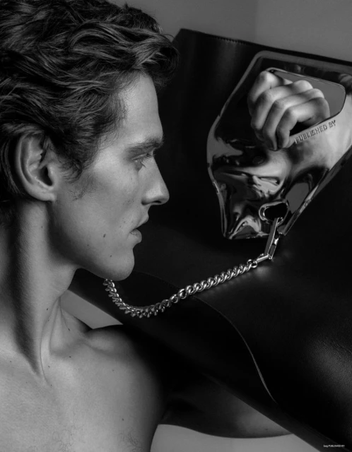 Monochrome image of a shirtless adult male in profile, with dark wavy hair, posing with a large black leather tote bag with a silver handle.