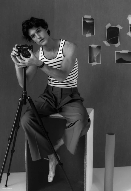Monochrome photo of a man seated with a camera on a tripod, striped tank top, neutral pants, in a studio, creating a sophisticated, artistic mood.