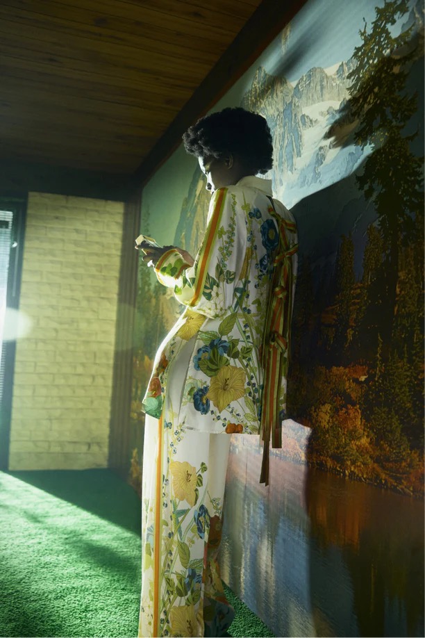 Woman in floral patterned pajama-style outfit holding phone indoors against a landscape-painted wall, fashion or interior photography.