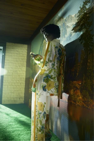 Woman in floral patterned pajama-style outfit holding phone indoors against a landscape-painted wall, fashion or interior photography.