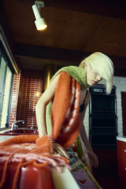 Young woman with blonde hair wears a fuzzy orange and green scarf indoors near a red countertop, kitchen cabinets, and a bright ceiling light.