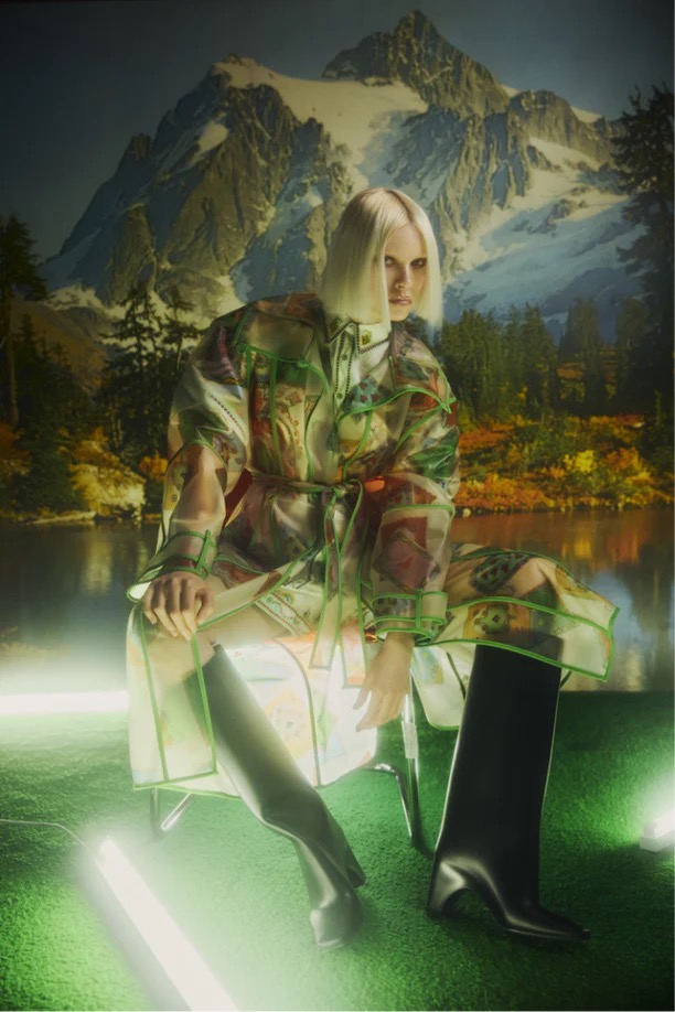 A fair-skinned person with short blonde hair poses in a patterned coat and black boots against a backdrop of mountains and a lake.
