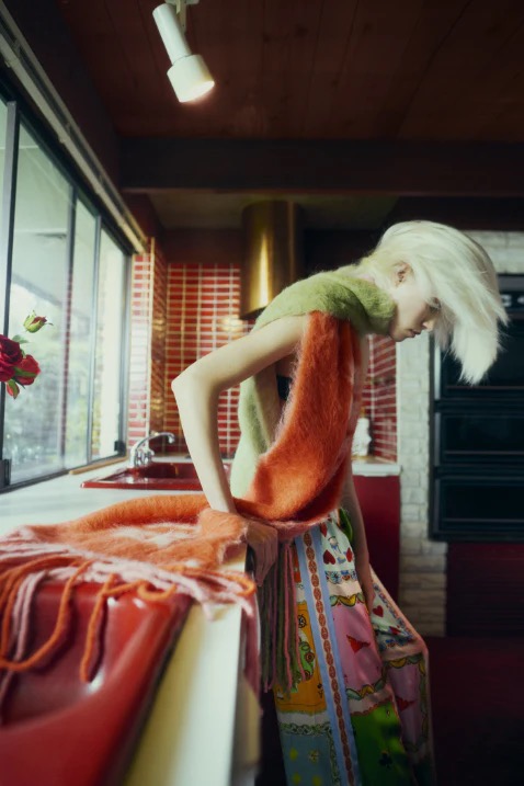 Woman with platinum blonde hair poses indoors wearing green top, orange scarf, and patterned skirt in a retro-style kitchen setting.