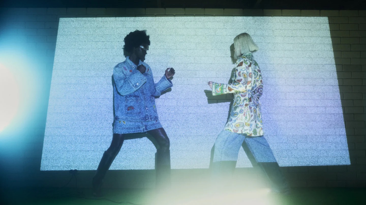 Two models face each other indoors under bright lights, wearing fashionable denim and paisley print clothing against a brick wall backdrop.