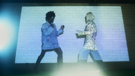 Two models standing in fighting poses against a brick wall projection for Alemais Pre Fall 24 Players campaign.