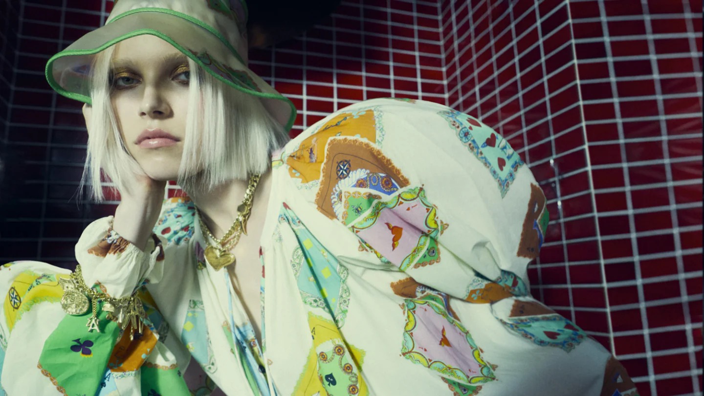 A woman with blonde hair and wearing a printed blouse and hat poses in front of a red tile wall for a fashion-themed portrait.
