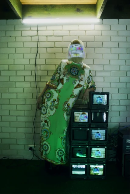 Artistic shot of a woman in patterned dress standing next to a stack of old TVs against a brick wall with bright neon lighting.