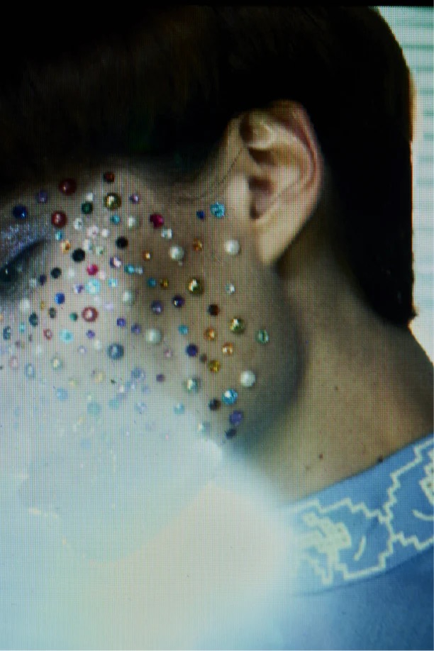 Close-up of a person with fair skin adorned with sparkling multi-colored rhinestones, focusing on face and neck, against a soft background.