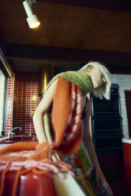 A woman poses in a brightly lit kitchen wearing a green and orange scarf and a patterned skirt in an editorial fashion image.
