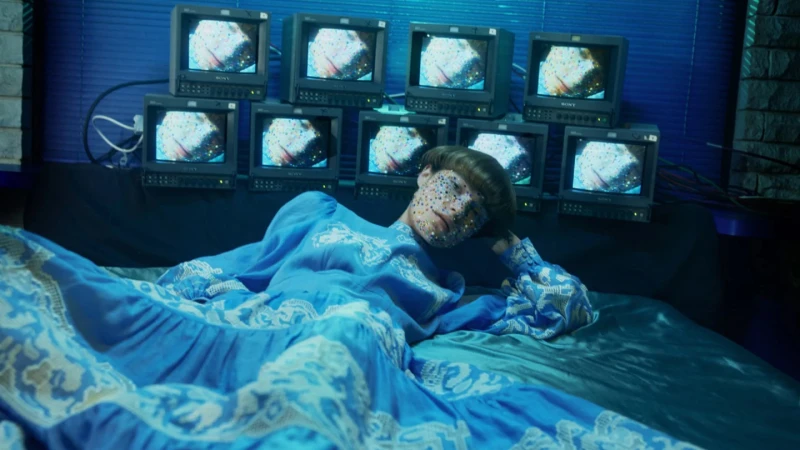 Woman in a patterned dress reclines on a bed with stacked Sony monitors displaying abstract images in a dark blue room with window blinds.