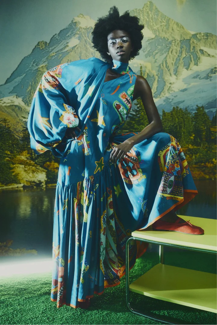 A black woman model poses in a long blue dress with graphic print against a mountain backdrop, with green foreground and yellow table.
