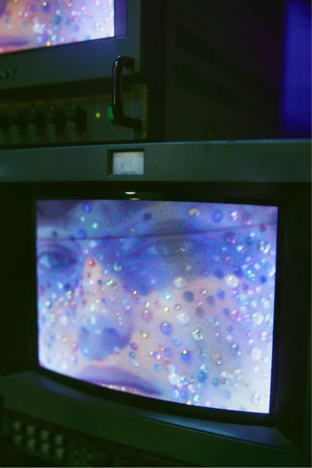 Television screens display a face covered in glitter in an indoor setting, with the focus on the screens and the displayed image.