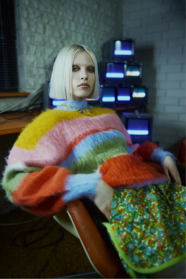 Woman with platinum blonde bob sits in chair wearing fuzzy striped sweater and printed skirt in a room with old televisions.