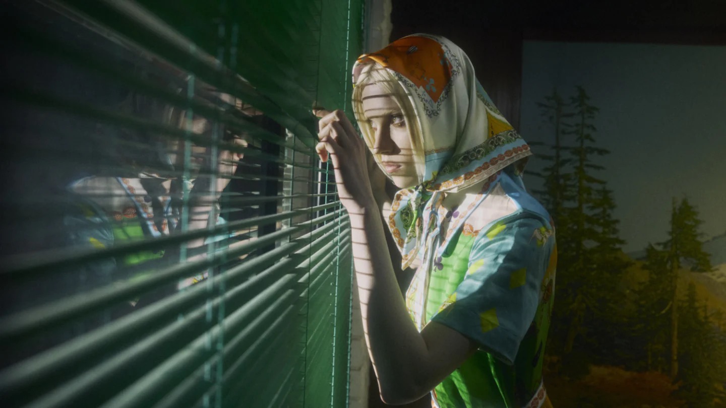 A young adult female is shown in an eye-level medium shot, looking through a window with green blinds and a silk patterned headscarf.