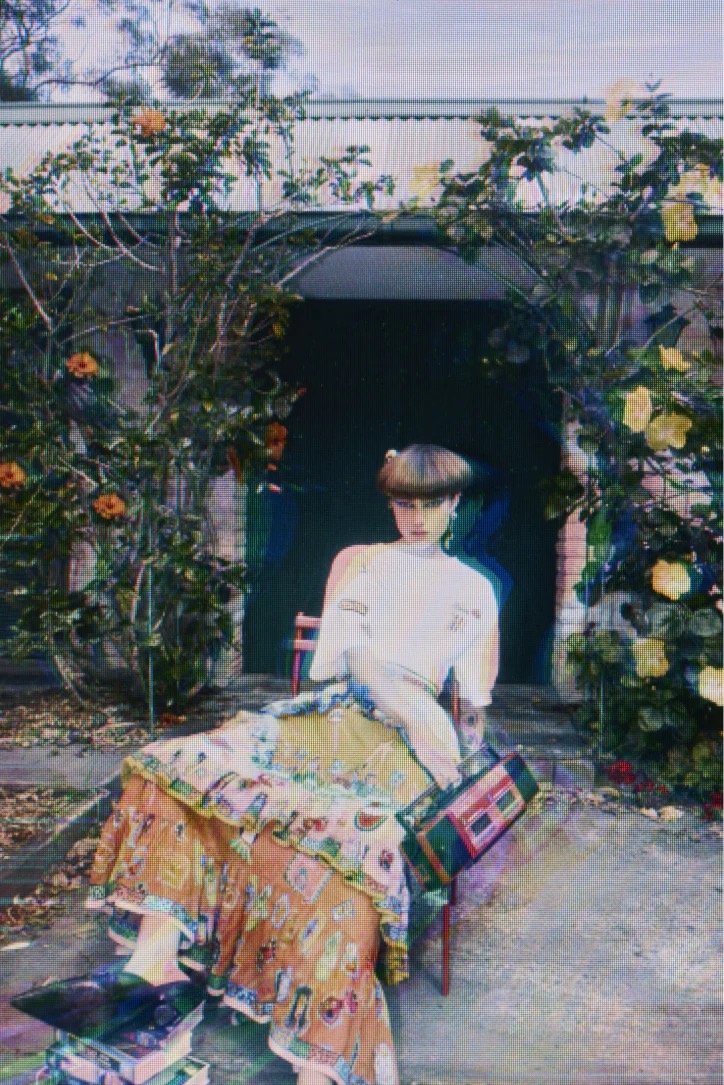 Woman in patterned dress sitting on chair surrounded by flowers and greenery, holding bag, in outdoor setting with a vintage aesthetic.