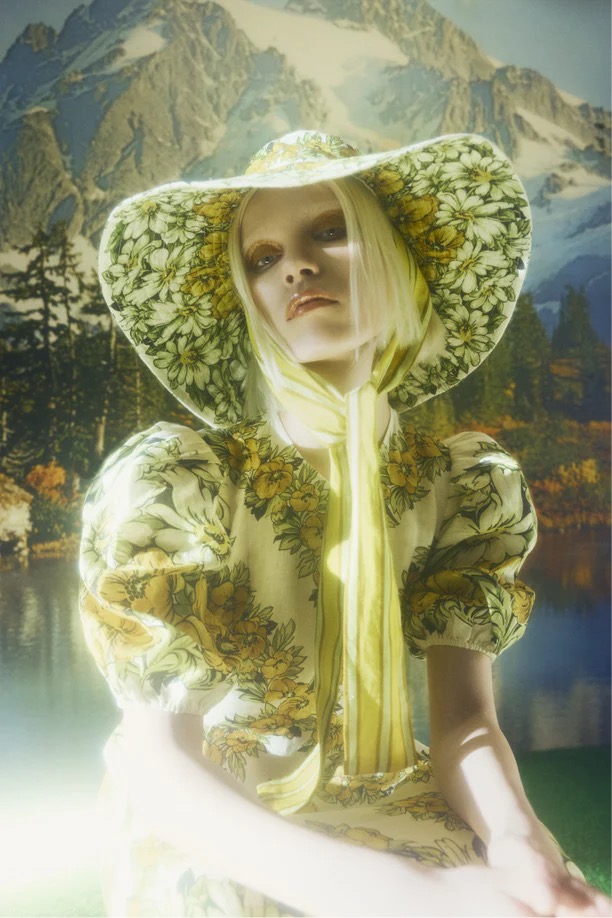 Adult female fashion model in wide-brimmed hat with a floral dress posing in front of a nature backdrop.