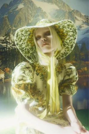 Adult female fashion model in wide-brimmed hat with a floral dress posing in front of a nature backdrop.