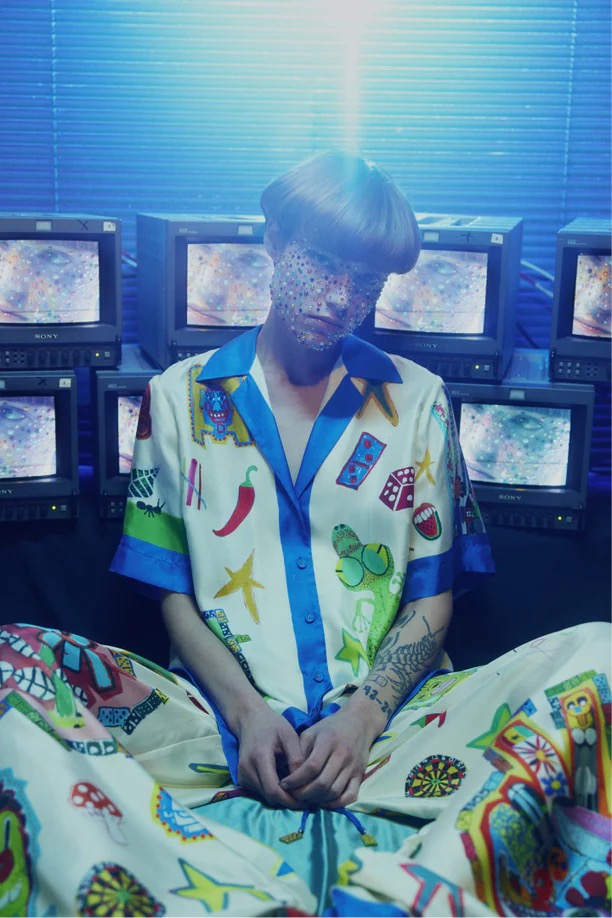 Adult in patterned pajamas sits against a wall of vintage televisions in a blue-lit room, presenting a fashion-forward, retro aesthetic.
