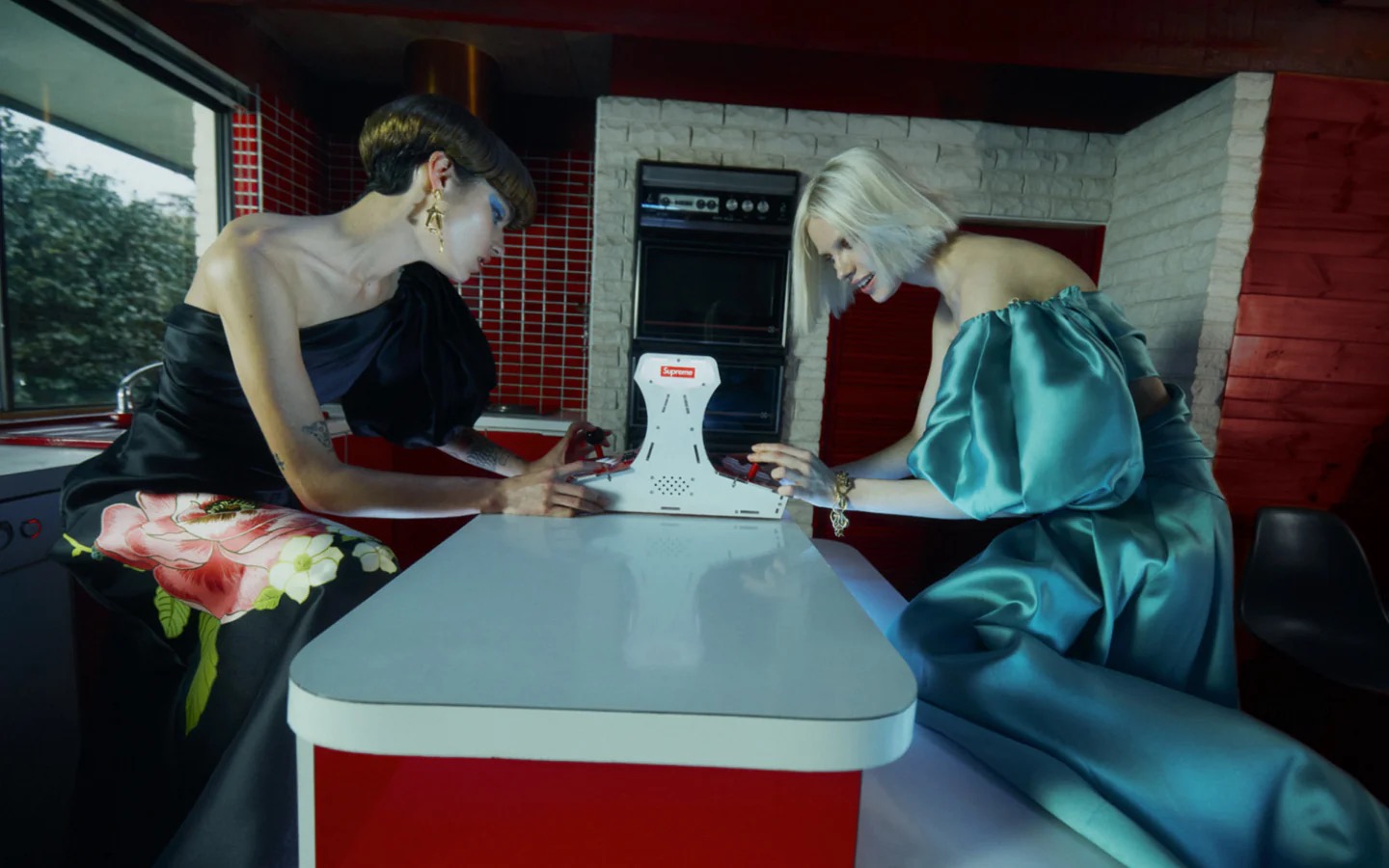 Two models in satin gowns pose at a table with a Supreme gaming machine in a retro-style kitchen with red and white accents.