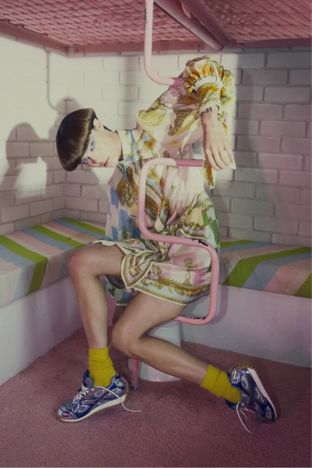 Person in patterned shirt and shorts sitting in a pink chair with yellow socks and sneakers in an indoor setting with striped walls.