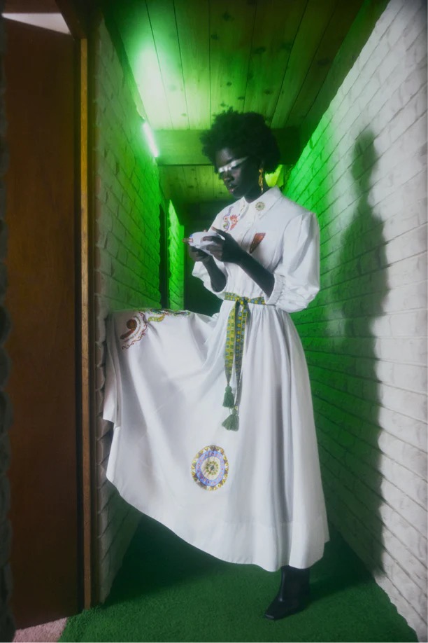 An adult female poses in a white dress with green accents and glasses in an indoor hallway with brick and wood-paneled walls.