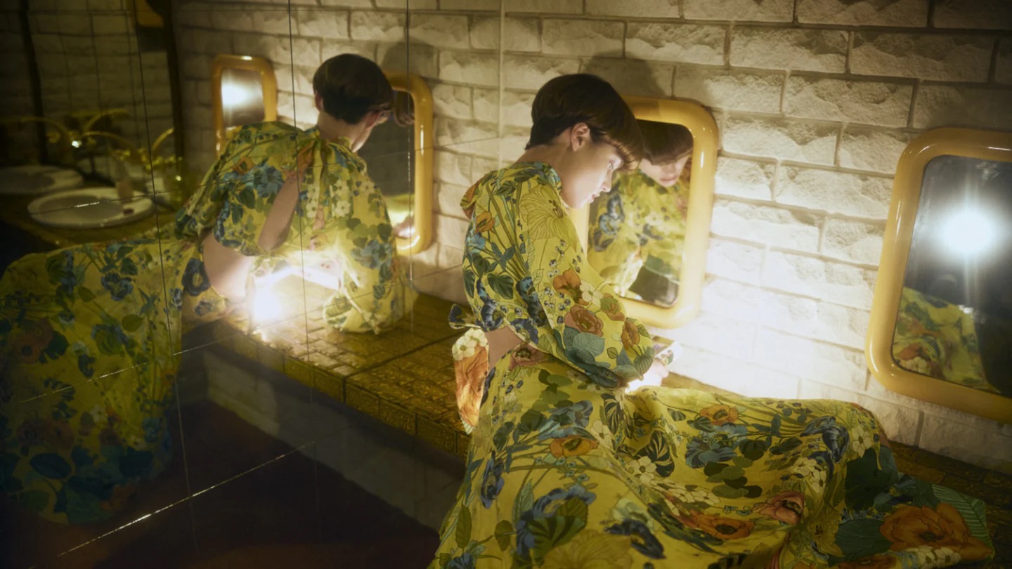Woman in floral dress sits at bathroom vanity with mirrors. She appears in multiple mirror reflections in an indoor environment.