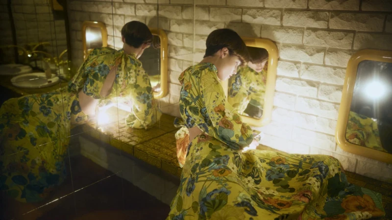 Woman in floral dress sits at bathroom vanity with mirrors.  She appears in multiple mirror reflections in an indoor environment.