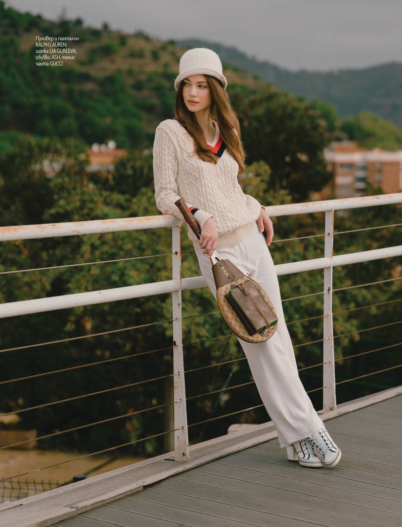 Indoor fashion shot of a woman in cream-colored knit sweater and white pants holding a tennis racket, with a mountain backdrop.