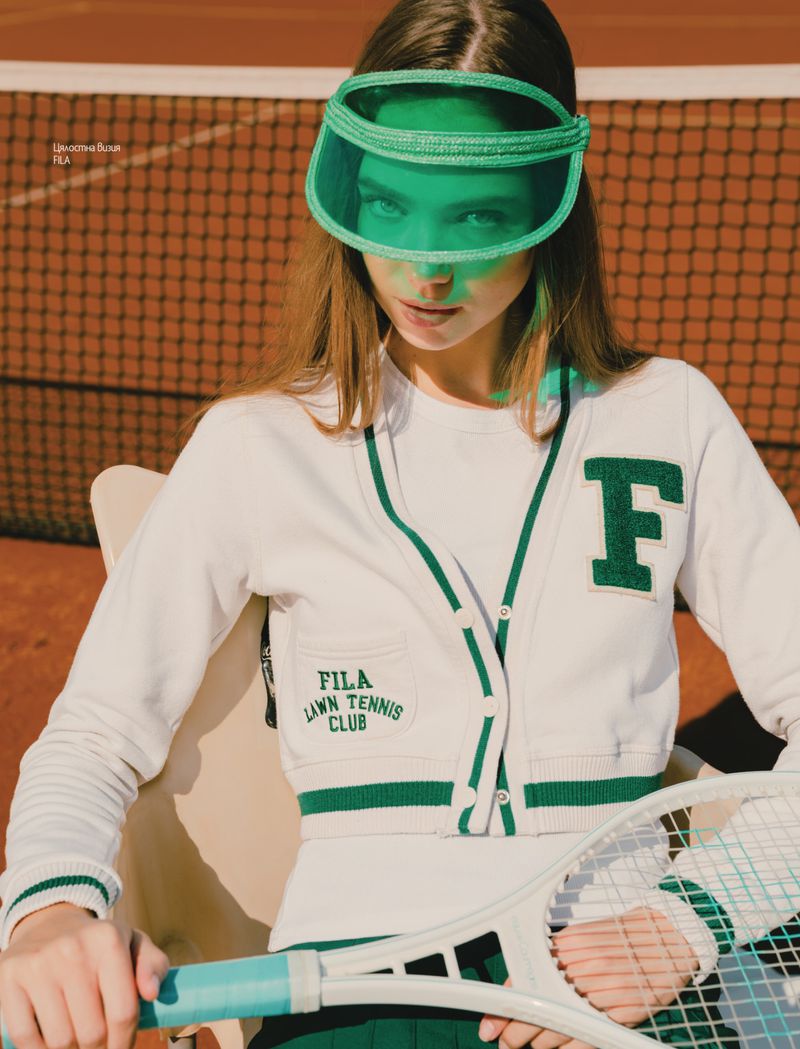 A woman poses on a tennis court in a white tennis cardigan with green trim, a green visor, and tennis racket during daytime.