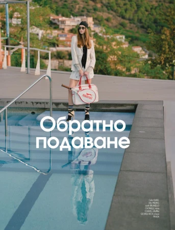 Fashionable woman with sunglasses poses near a pool with a tennis bag and racket, featuring a backdrop of architecture and landscape.