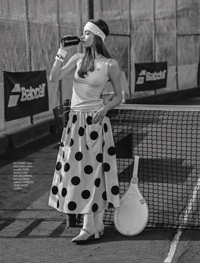 Black and white shot of a female tennis player in a polka dot skirt drinking from a container next to a tennis net and racket.