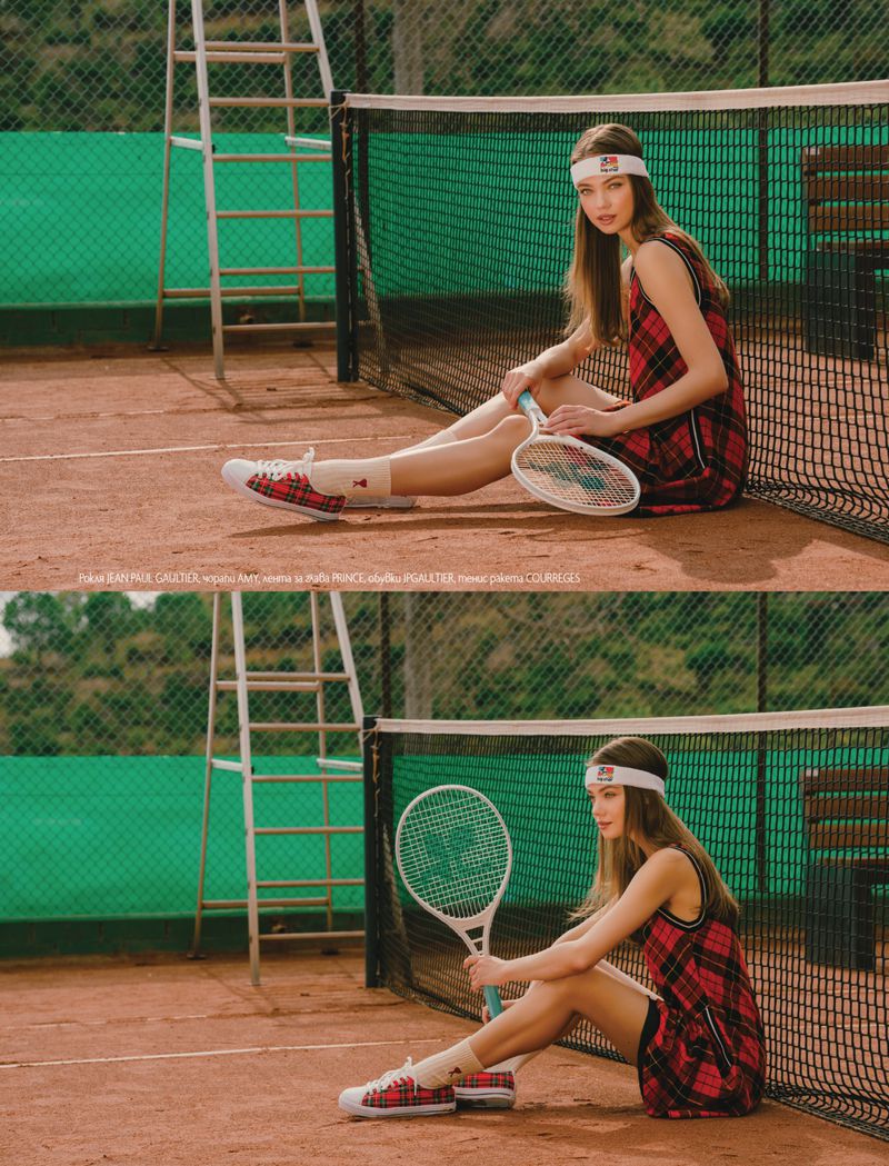 A young female model wearing a red plaid dress and white headband holds a tennis racket while seated on a clay tennis court.