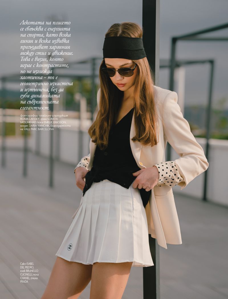 Woman in fashion editorial poses in a white pleated skirt with beige jacket and black top, wearing sunglasses and a headband outdoors.