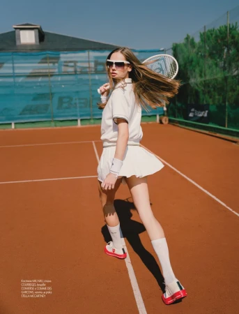 A woman on a tennis court holds a racquet, wearing a white tennis outfit and sunglasses, posing with a tennis court background.