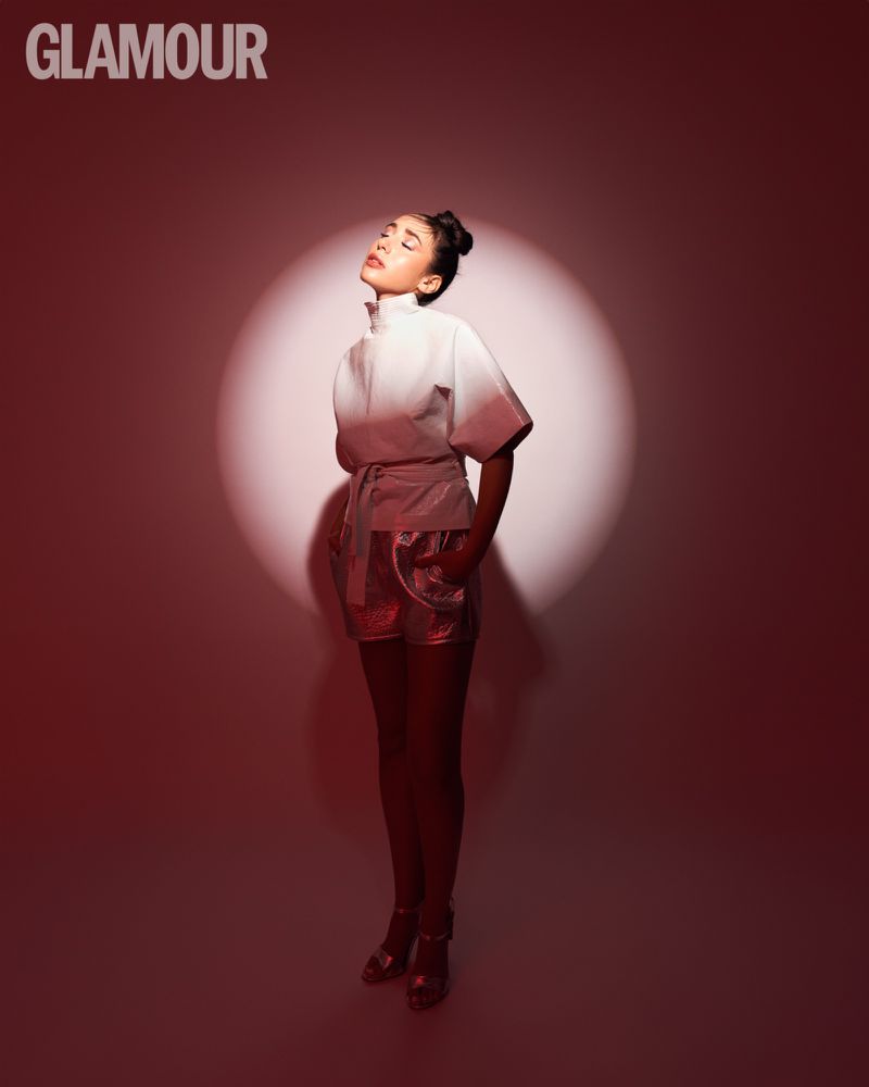 Woman in trendy outfit posing in photo studio with white top and shiny shorts, illuminated by a spotlight with red lighting in background.