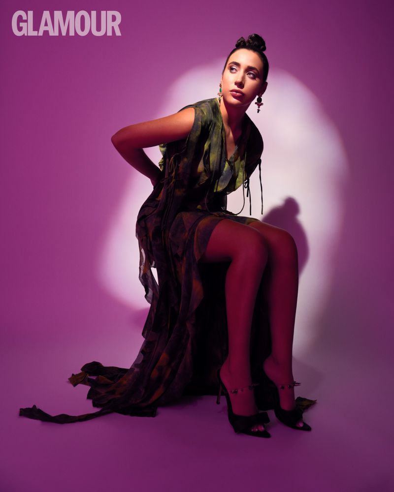 Portrait of a woman in a printed dress sitting in a photo studio with a purple background. She is wearing black high heels.