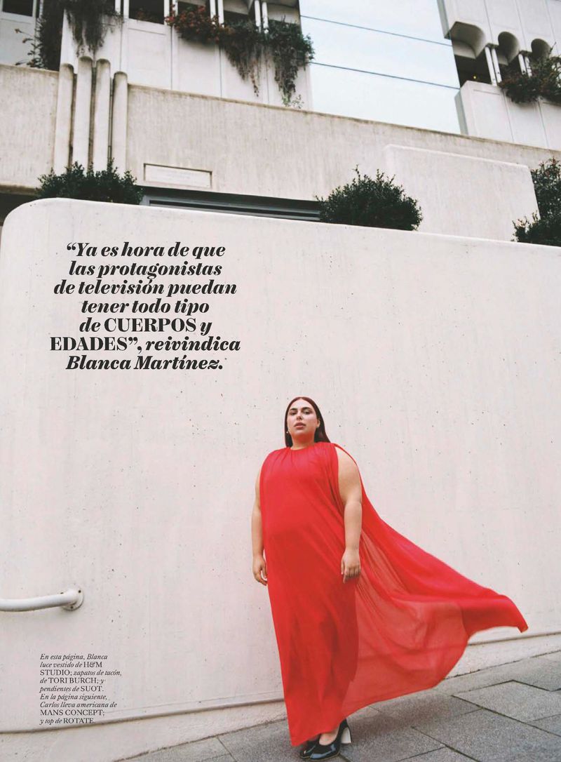 Adult woman in a red dress poses against a white wall with architectural details, under outdoor lighting in an editorial fashion setting.