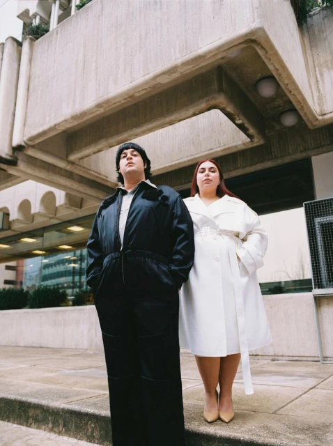 Two plus-size adult women pose in front of concrete architecture, wearing a black jumpsuit and white trench coat, respectively, under an overcast sky.
