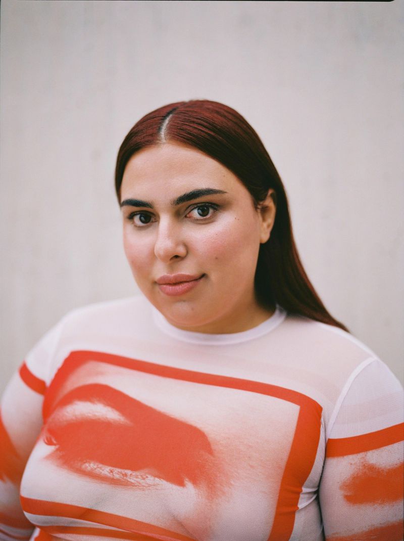 Close-up portrait of a brunette adult female in a white patterned top against a plain wall in a studio. Beauty shot.