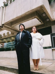 Two models posing outdoors beneath a concrete brutalist structure for Vogue Espana, wearing black jumpsuit and white coat