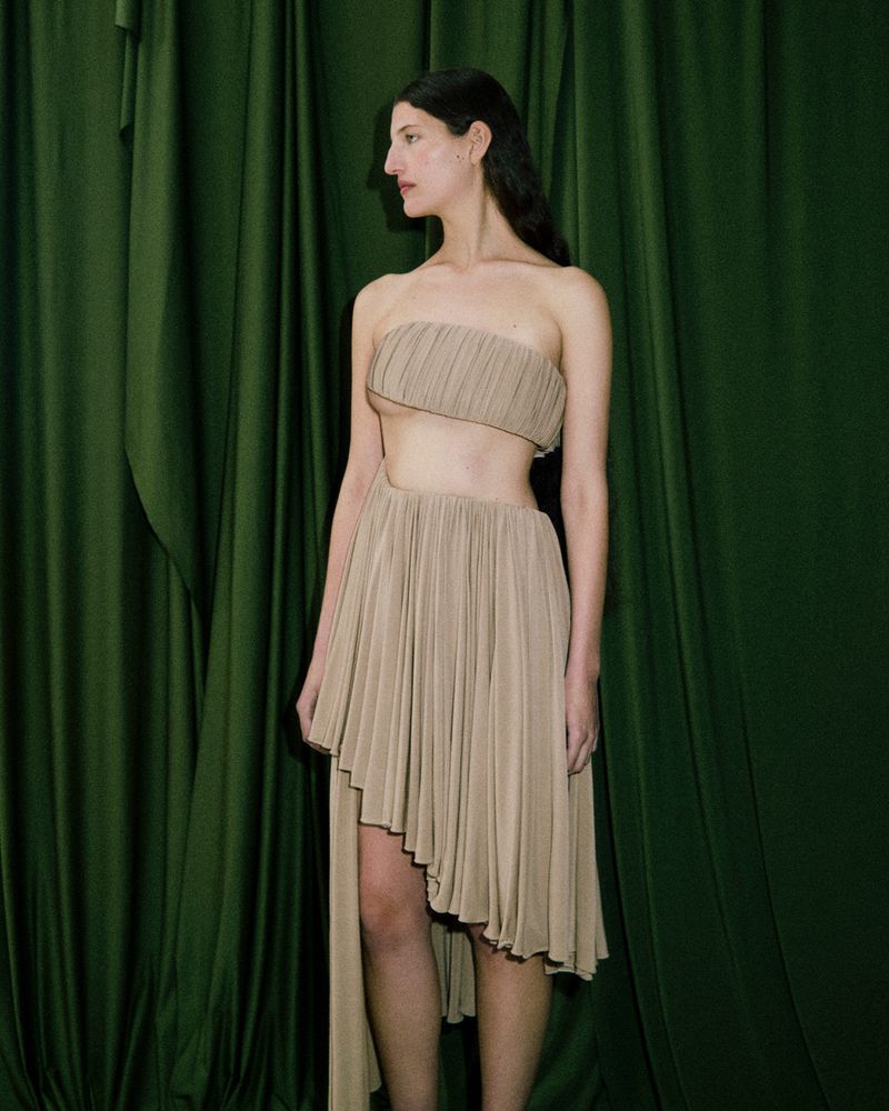 Model wearing a pleated beige dress standing against dark green draped fabric background