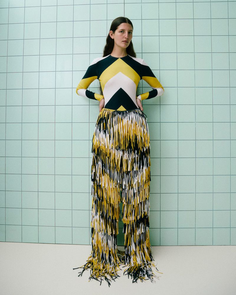 A woman poses indoors wearing a yellow, white, and black geometric pattern top, and yellow, white, and black fringe pants.